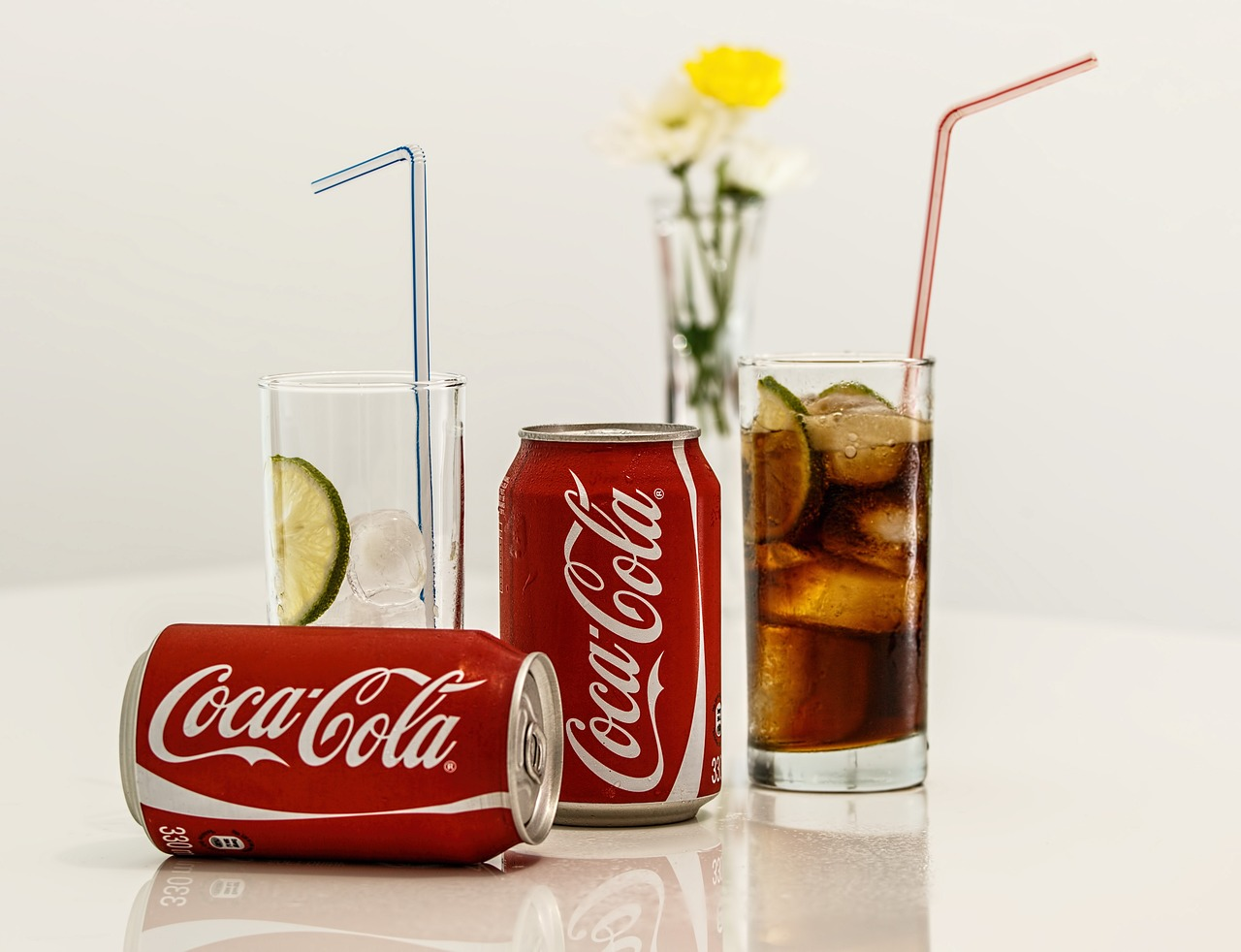 Two Coca-Cola cans, a glass of lemon water with ice and a glass of Coke sitting on a platform