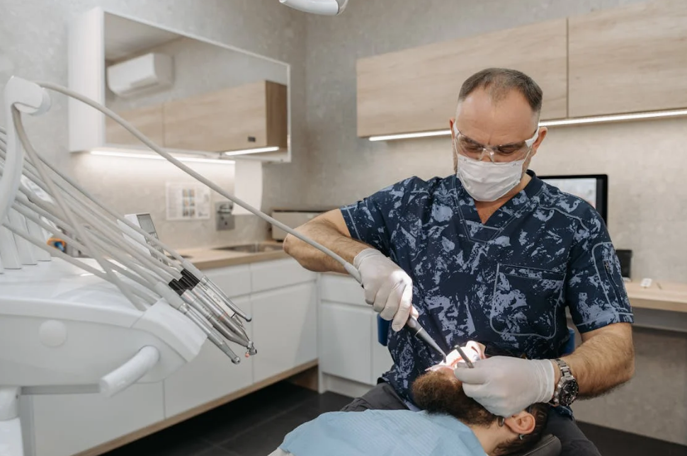 A dentist in protective gear performing a dental procedure on a bearded patient